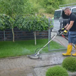 Pracownik w żółtych gumowcach czyści kostkę brukową za pomocą profesjonalnej myjki ciśnieniowej z obrotową głowicą, na tle srebrnego busa i zieleni.