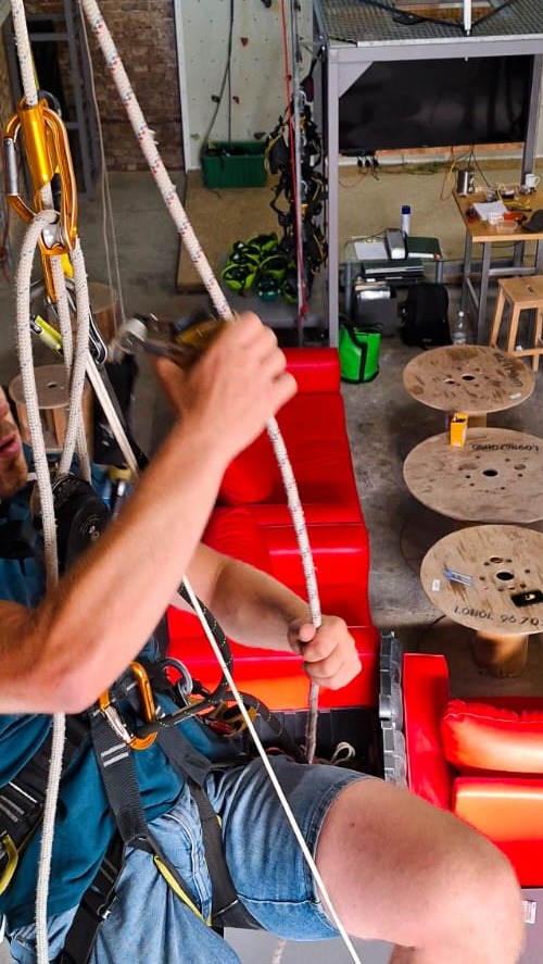 Mężczyzna w uprzęży alpinistycznej, przygotowujący się do pracy na wysokości, trzyma grubą linę asekuracyjną, w tle widoczne elementy wyposażenia wspinaczkowego i wypoczynkowa strefa biura...