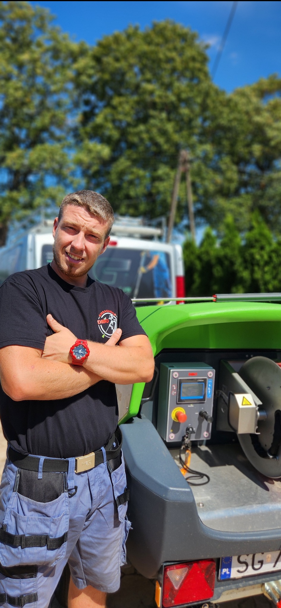 Uśmiechnięty mężczyzna w roboczym stroju, z założonymi rękami, stoi obok specjalistycznej maszyny z panelem sterowania, prawdopodobnie do czyszczenia powierzchni, z zaparkowanym vanem w tle.
