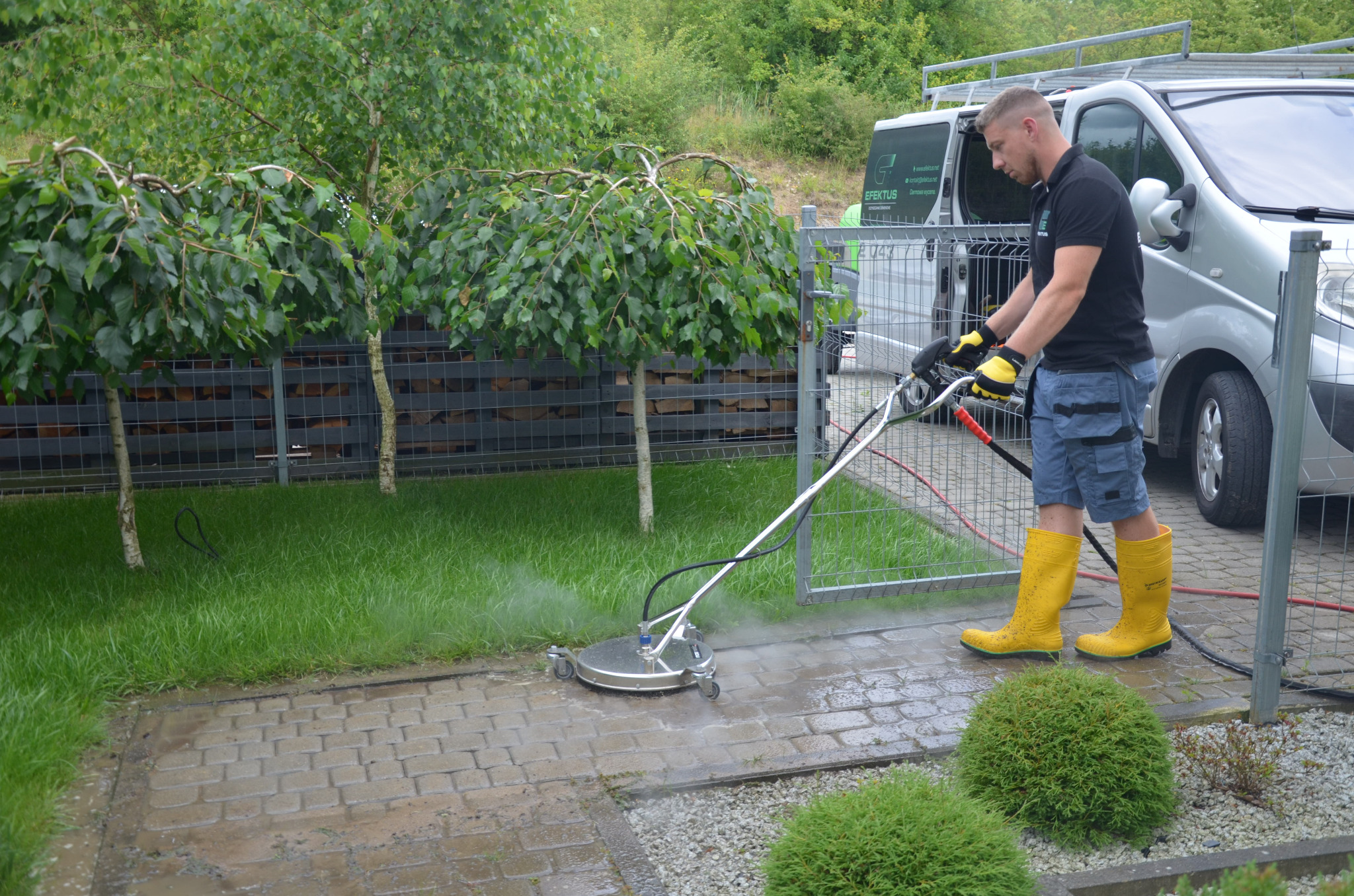 Pracownik w żółtych gumowcach czyści kostkę brukową za pomocą profesjonalnej myjki ciśnieniowej z obrotową głowicą, na tle srebrnego busa i zieleni.