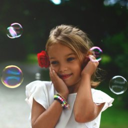 Belisana - Uśmiechnięta dziewczynka z czerwoną różą we włosach dotyka dłońmi policzków, otoczona unoszącymi się bańkami mydlanymi, w tle zieleń parku.