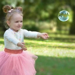 Belisana - Dziewczynka w różowej tiulowej spódniczce próbuje złapać bańkę mydlaną na tle zielonego parku.