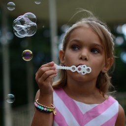 Belisana - Dziewczynka w sukience w różowe paski dmucha bańki mydlane przez plastikową foremkę, w tle zielona roślinność.