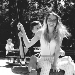 Belisana - Czarno-białe zdjęcie uśmiechniętej kobiety w okularach w kształcie serc siedzącej na huśtawce z mężczyzną w parku, w tle widoczna karuzela z dziećmi.