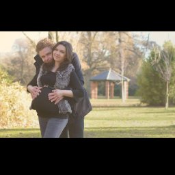 Belisana - Przyszła mama w futrzanej kamizelce, obejmowana przez partnera na tle parku z altaną.