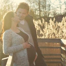 Belisana - Kobieta w ciąży, w jasnym kardiganie, stoi na drewnianym mostku z partnerem, oboje patrzą na siebie, w tle wysoka trawa i zachodzące słońce.