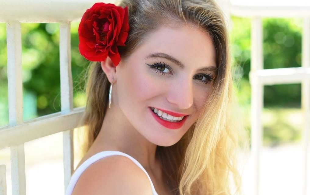 Portret uśmiechniętej blondynki z czerwoną różą we włosach, oparta o białą balustradę, z zielonym tłem.