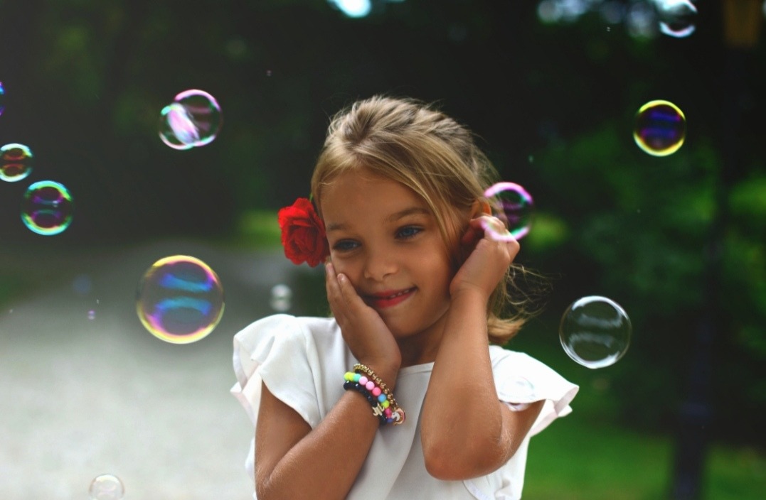 Uśmiechnięta dziewczynka z czerwoną różą we włosach dotyka dłońmi policzków, otoczona unoszącymi się bańkami mydlanymi, w tle zieleń parku.