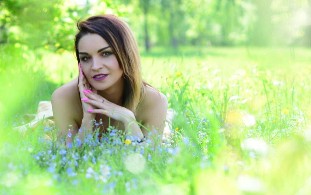 Portret kobiety leżącej na łące w plenerze, z dłońmi ułożonymi pod brodą, z widocznymi niebieskimi kwiatami i rozproszonym światłem słonecznym.