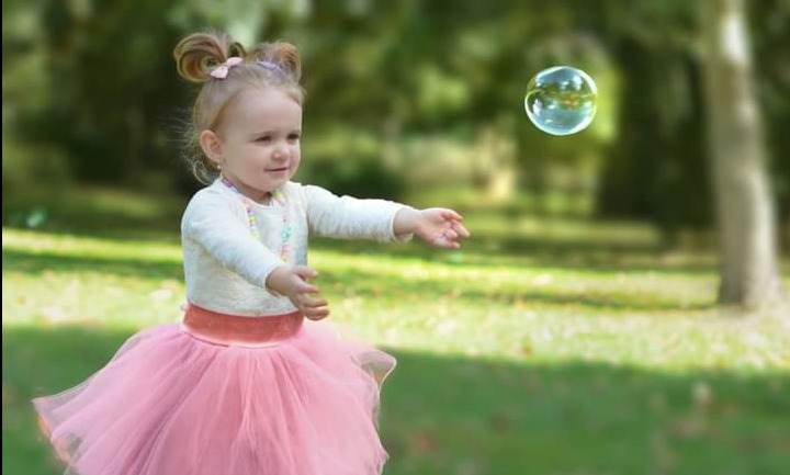 Dziewczynka w różowej tiulowej spódniczce próbuje złapać bańkę mydlaną na tle zielonego parku.