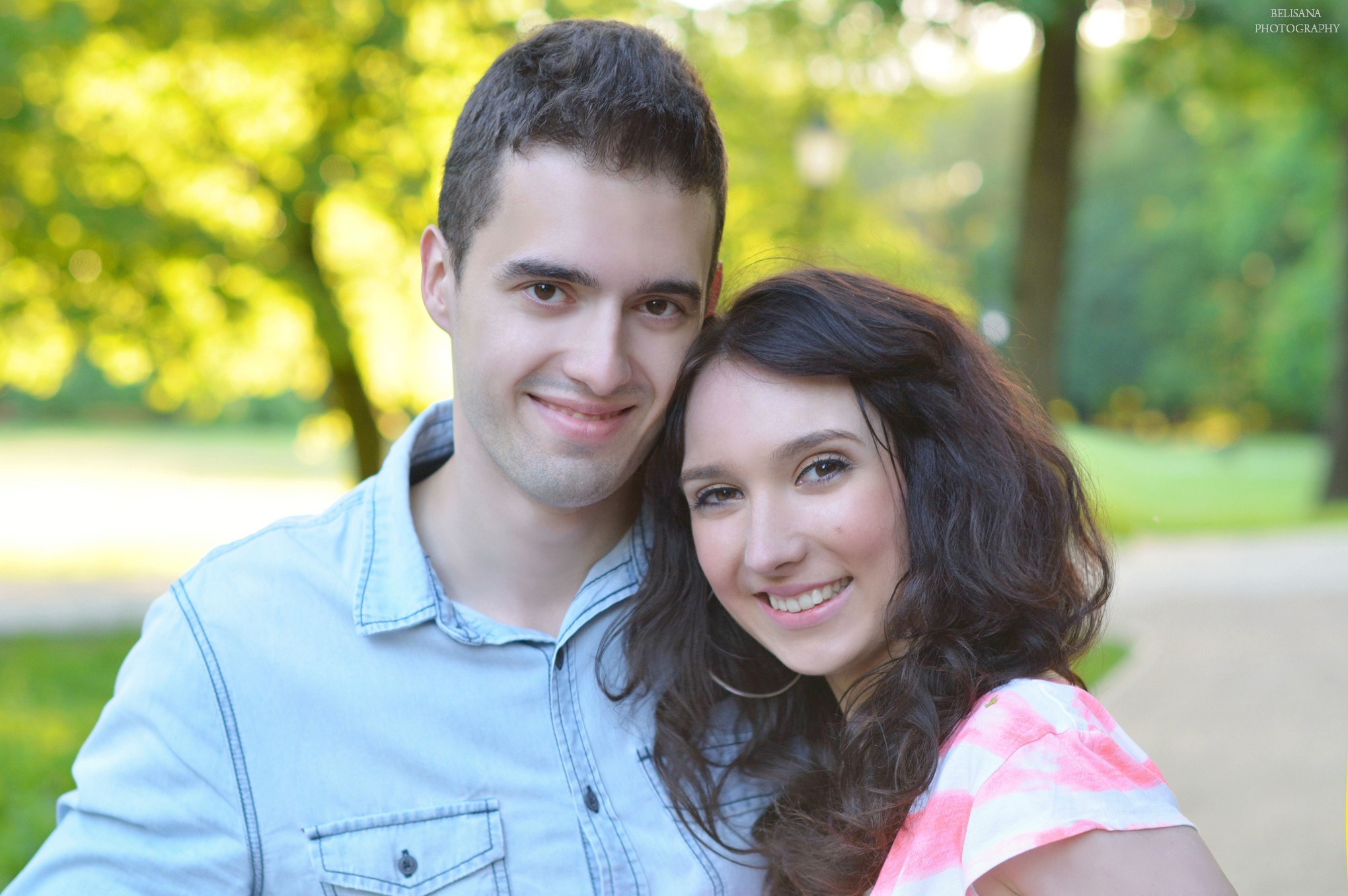 Uśmiechnięta para wtulona w siebie na tle zielonego parku, mężczyzna w jasnoniebieskiej koszuli, kobieta w różowo-białej bluzce, delikatne rozmycie tła.