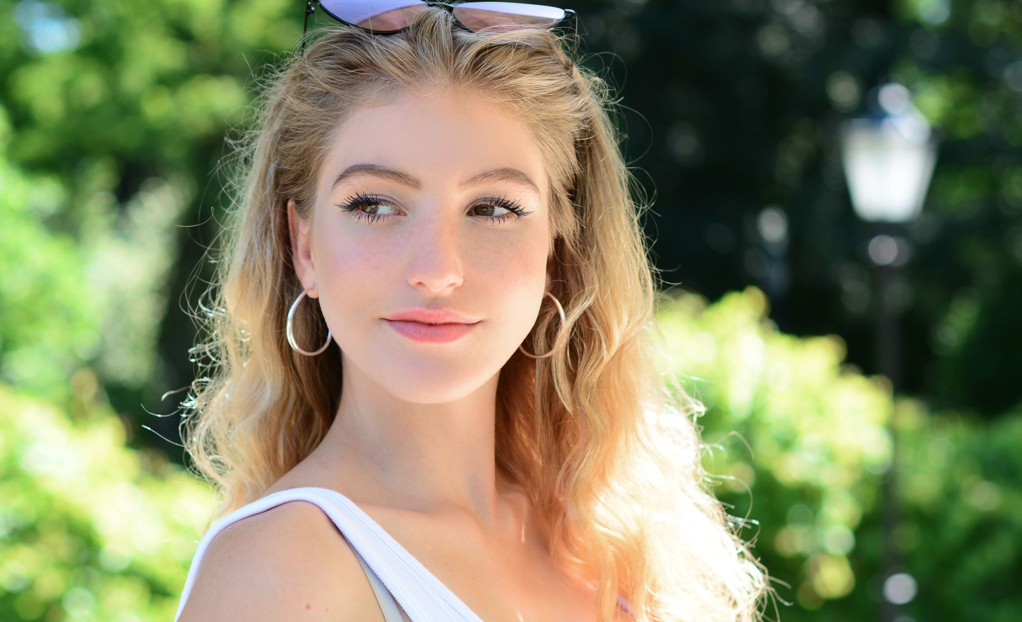 Portret młodej kobiety z blond włosami, dużymi kolczykami kołami i okularami na głowie, spoglądającej w bok na tle zieleni parkowej.