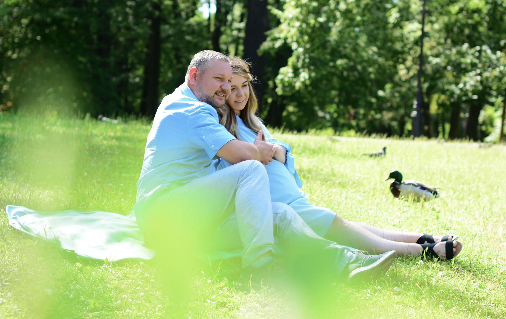 Para siedząca na kocu w parku, mężczyzna obejmuje kobietę, w tle kaczki na trawie.