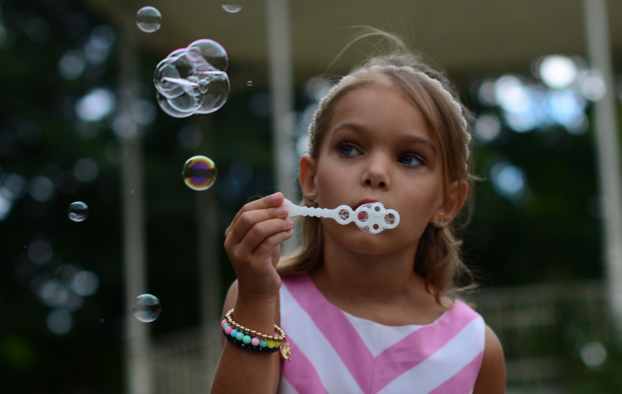 Dziewczynka w sukience w różowe paski dmucha bańki mydlane przez plastikową foremkę, w tle zielona roślinność.