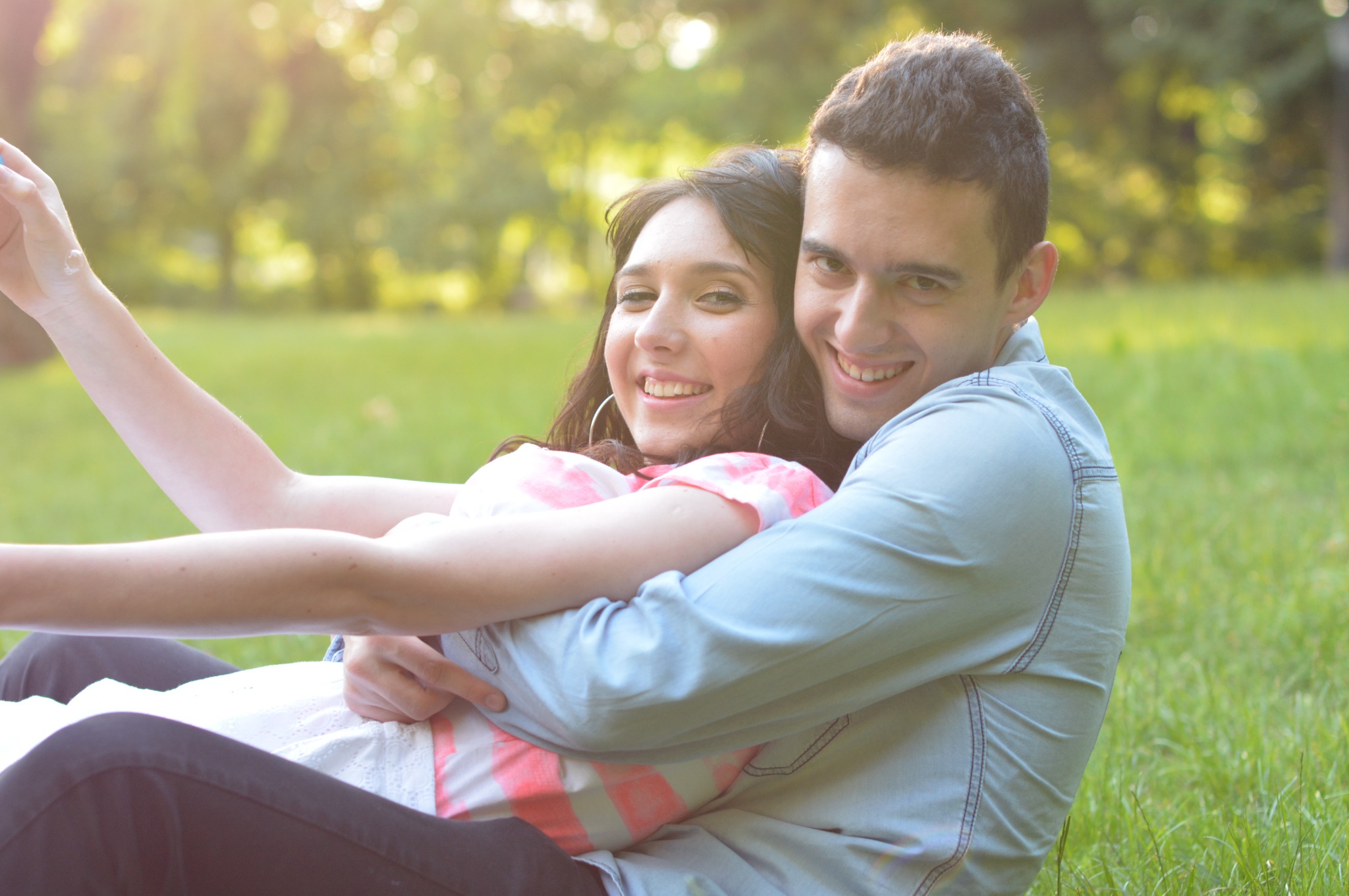 Uśmiechnięta para narzeczonych siedzi na trawie w parku, obejmując się w promieniach zachodzącego słońca, kobieta w sukience w różowe paski i kolczykami kołami, mężczyzna w jeansowej koszuli.