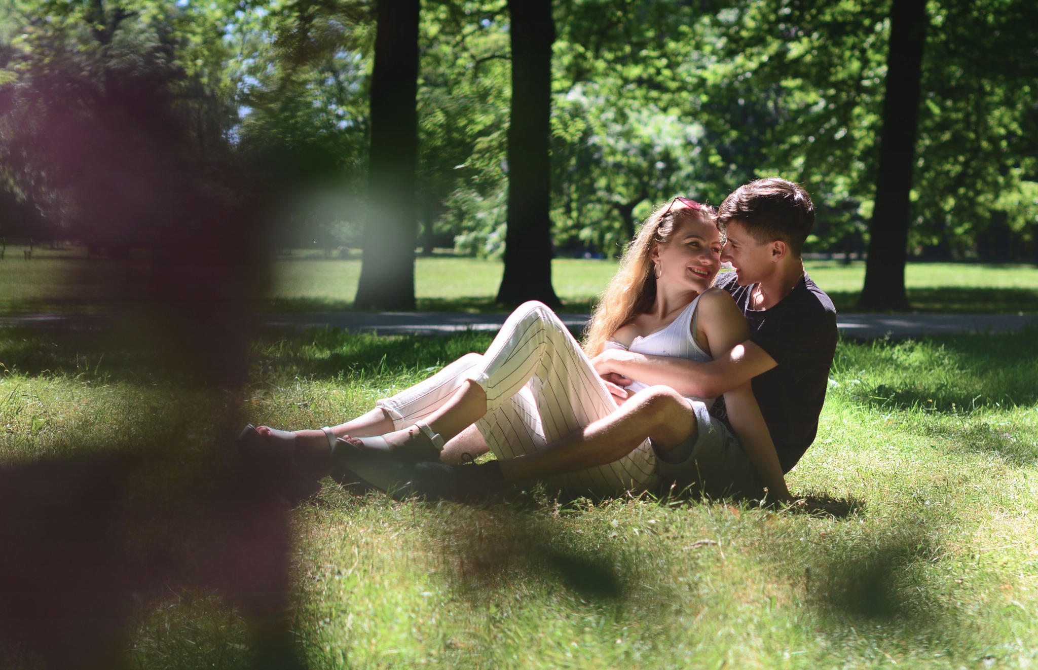 Młoda para siedzi na trawie w parku, obejmując się i patrząc sobie w oczy, z promieniami słońca przebijającymi przez drzewa w tle.