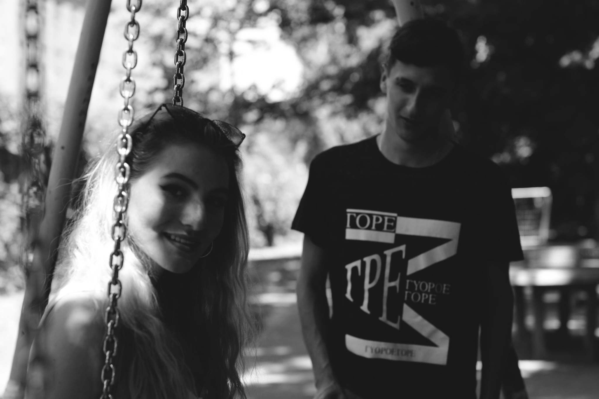 Czarno-białe zdjęcie uśmiechniętej kobiety na huśtawce łańcuchowej, z mężczyzną w tle, oboje wyglądają na młodych, swobodna atmosfera.