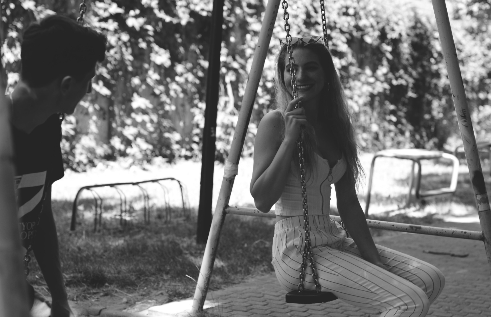 Czarno-białe zdjęcie uśmiechniętej kobiety w paski siedzącej na huśtawce, z okularami na głowie, w tle zarośla i fragment chłopca z lewej strony.