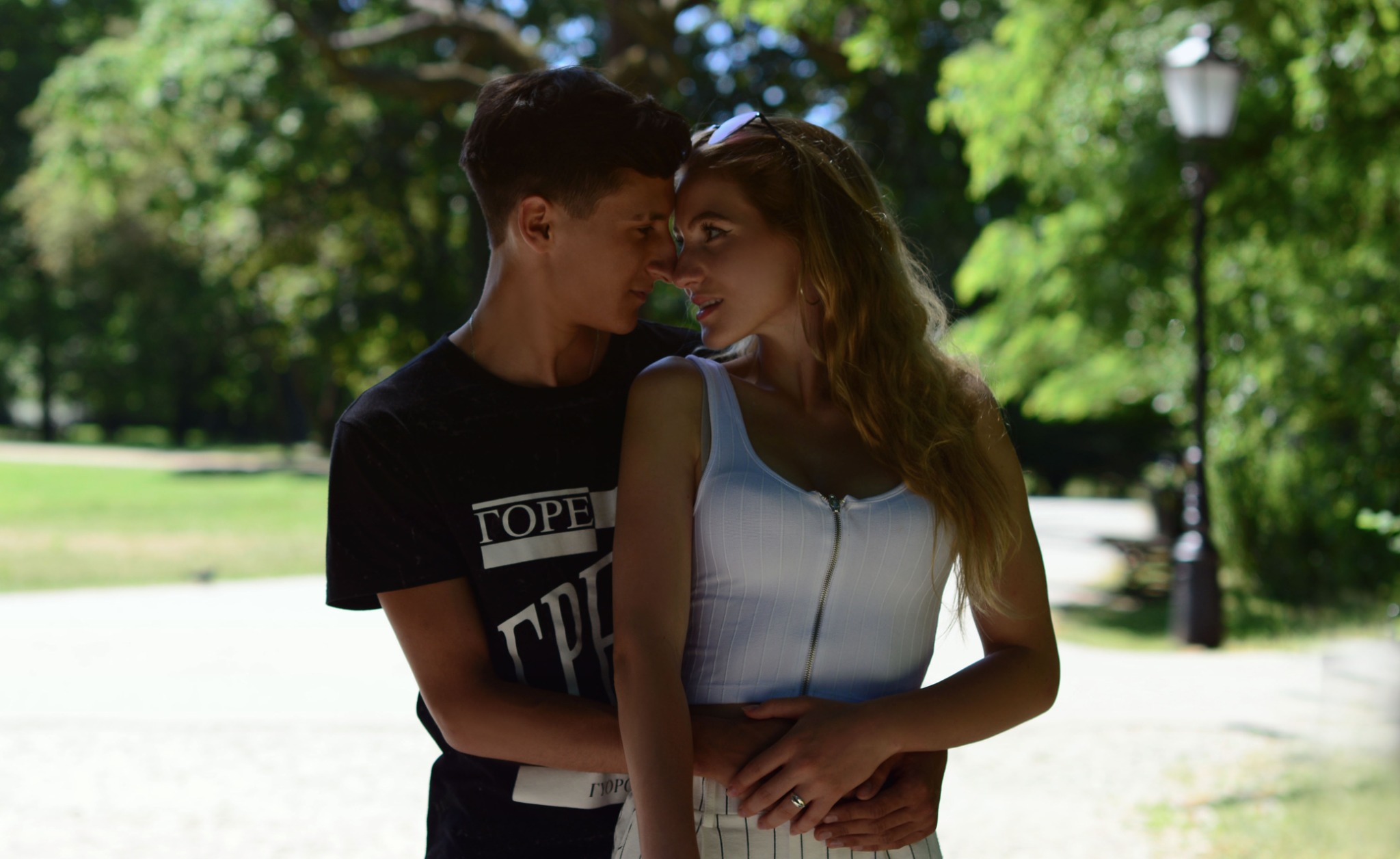 Młoda para, chłopak w czarnej koszulce z napisem, obejmuje dziewczynę w białym topie na tle parku, zbliżają się twarzami.