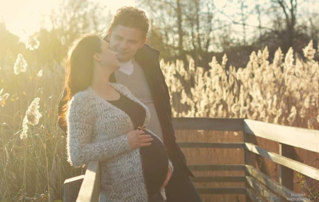 Kobieta w ciąży, w jasnym kardiganie, stoi na drewnianym mostku z partnerem, oboje patrzą na siebie, w tle wysoka trawa i zachodzące słońce.