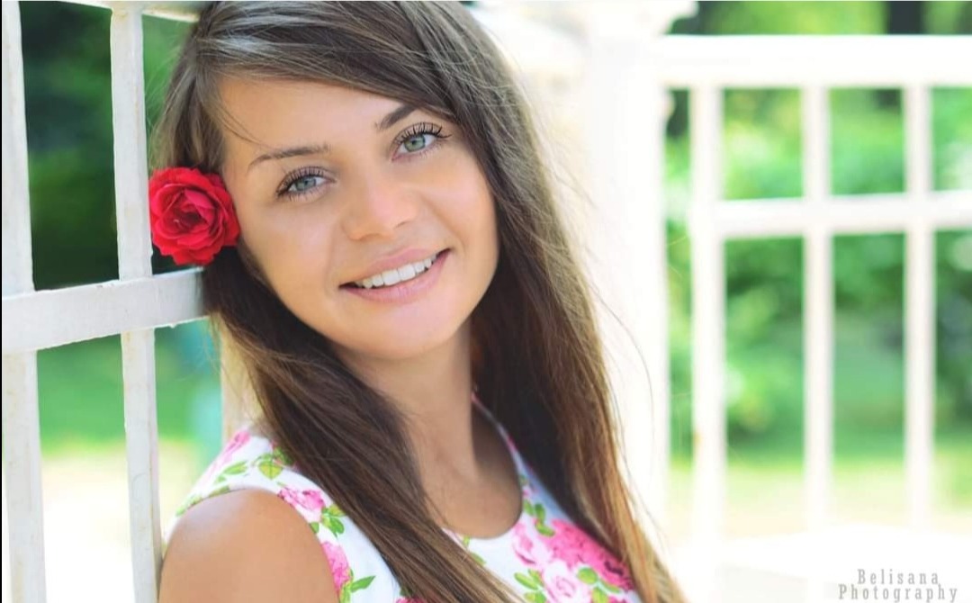 Portret uśmiechniętej brunetki z czerwoną różą we włosach, opierającej się o białą, drewnianą konstrukcję na tle zieleni.