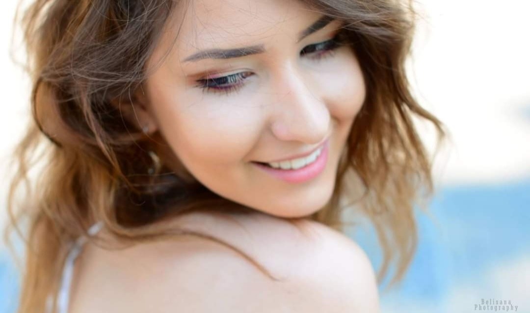 Portret uśmiechniętej kobiety z delikatnym makijażem, sfotografowanej z profilu w Łodzi, z widocznym fragmentem ramienia i rozpuszczonymi włosami.