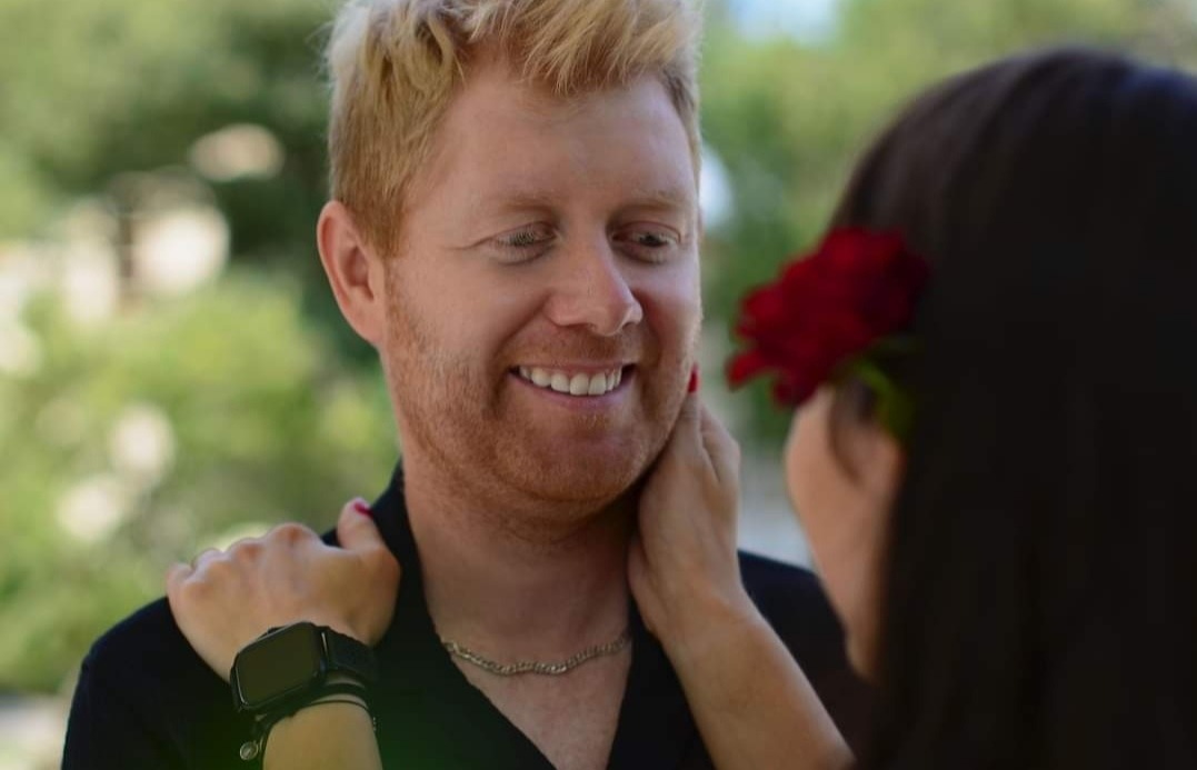 Uśmiechnięty rudowłosy mężczyzna z łańcuszkiem, dotykany przez kobietę z czerwoną różą we włosach, na tle zieleni.