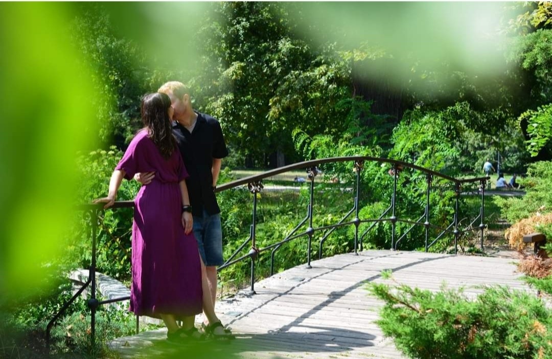 Para całująca się na drewnianym mostku w parku, kobieta w długiej fioletowej sukience, mężczyzna w czarnej koszulce i dżinsowych szortach, otoczeni zielenią drzew i krzewów.