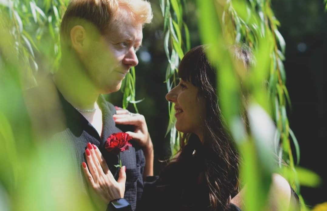 Zakochana para w plenerze, kobieta z ciemnymi włosami opiera ręce na piersi mężczyzny trzymającego czerwoną różę, uśmiechają się do siebie, widok przez gałęzie wierzby.