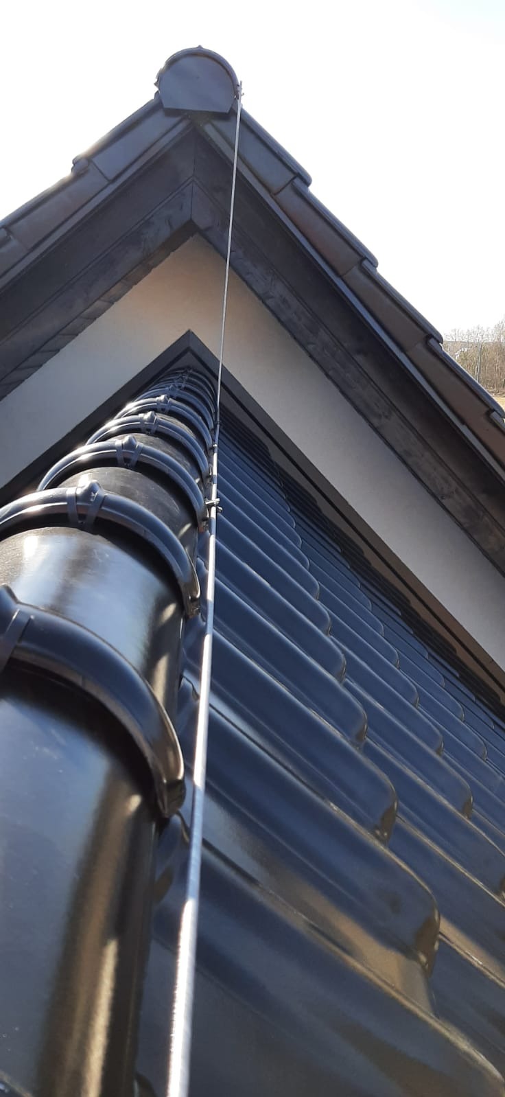 Błyszcząca, ciemna dachówka ceramiczna z zamocowanym przewodem odgromowym, widok z dołu na szczyt dachu.