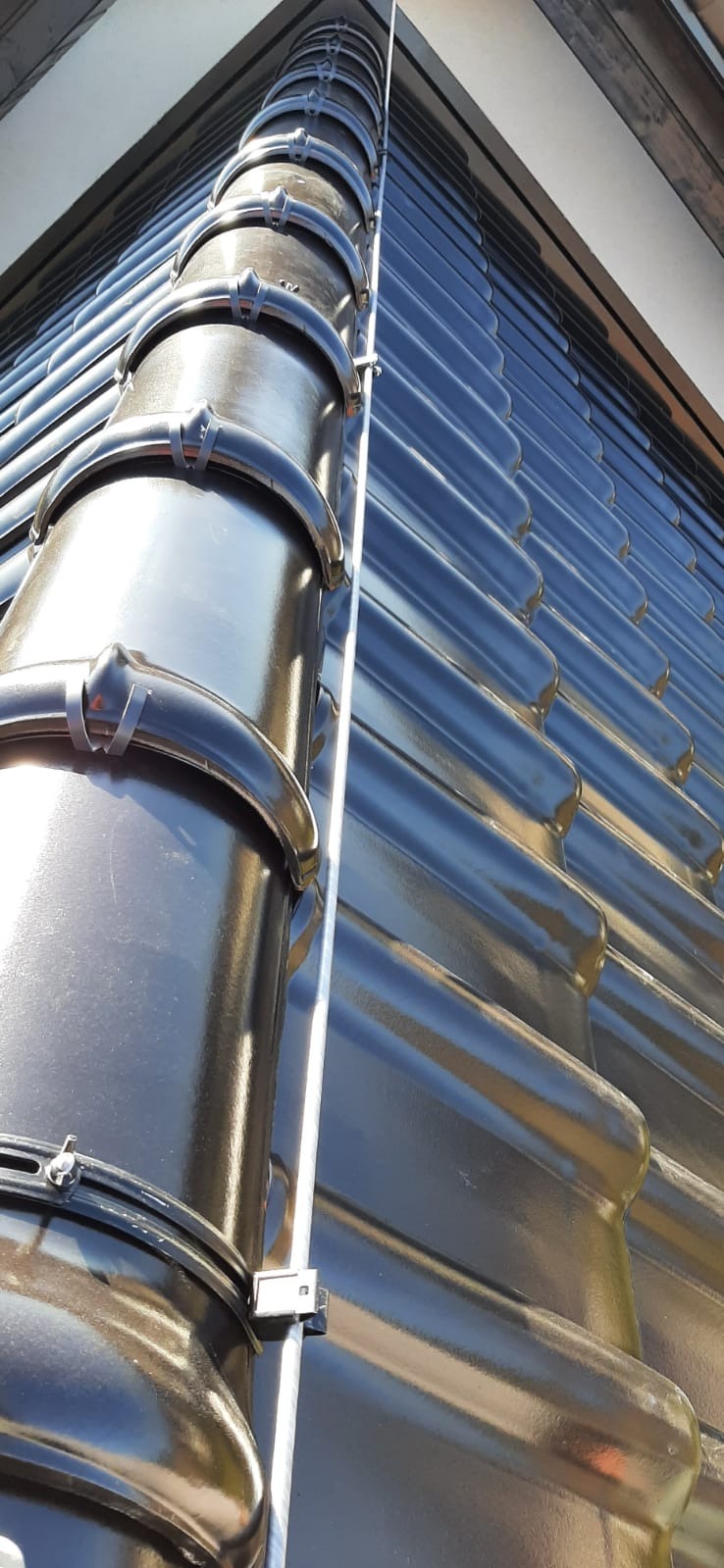 Błyszczący, czarny system rynnowy z przymocowanym przewodem odprowadzającym na dachu pokrytym ciemną dachówką ceramiczną, widoczne mocowania rur spustowych.