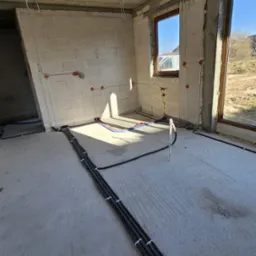 Surowy stan: instalacja elektryczna w peszlach i puszki na ścianach w nowym budynku, widok z góry na podłogę i okna. W tle widoczne otoczenie budynku.