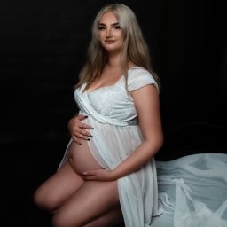 FRĄK STUDIO PHOTO Natalia Frąk - Kobieta w ciąży, na kolanach, w białej, zwiewnej sukience z koronkową górą, pozuje na czarnym tle, dłonie spoczywają na brzuchu, makijaż w odcieniach nude.