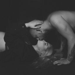 FRĄK STUDIO PHOTO Natalia Frąk - Czarno-białe zdjęcie pary: kobieta w koszuli i mężczyzna z tatuażem, leżący blisko siebie, dotykający się ustami.