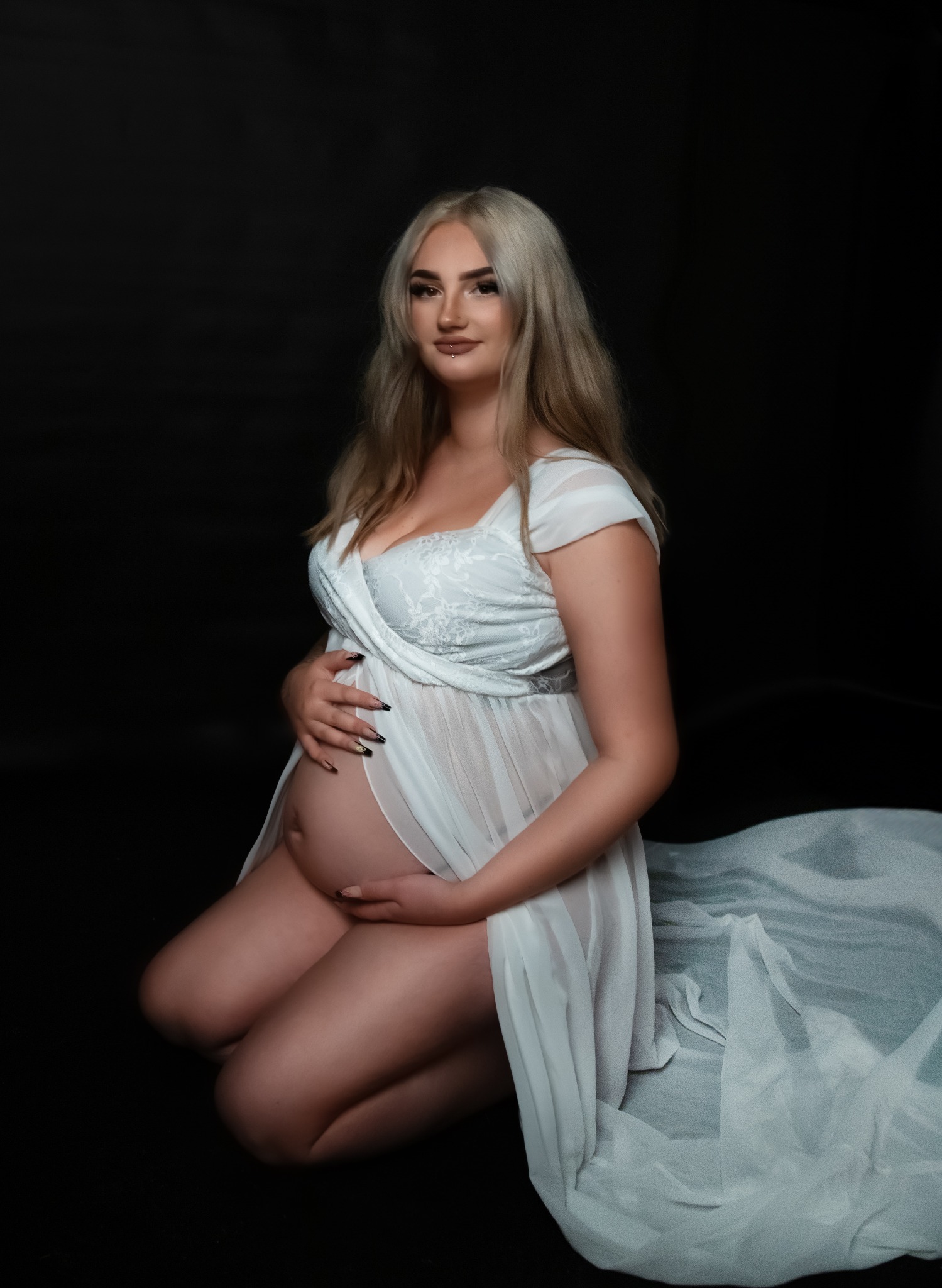 Kobieta w ciąży, na kolanach, w białej, zwiewnej sukience z koronkową górą, pozuje na czarnym tle, dłonie spoczywają na brzuchu, makijaż w odcieniach nude.