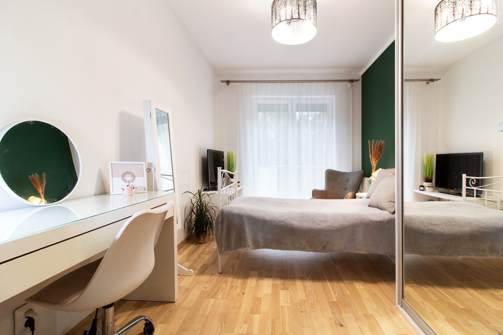 Jasna sypialnia z białym biurkiem, okrągłym lustrem z zielonym tłem, łóżkiem z szarą narzutą i przesuwną szafą z lustrem odbijającym część pokoju.