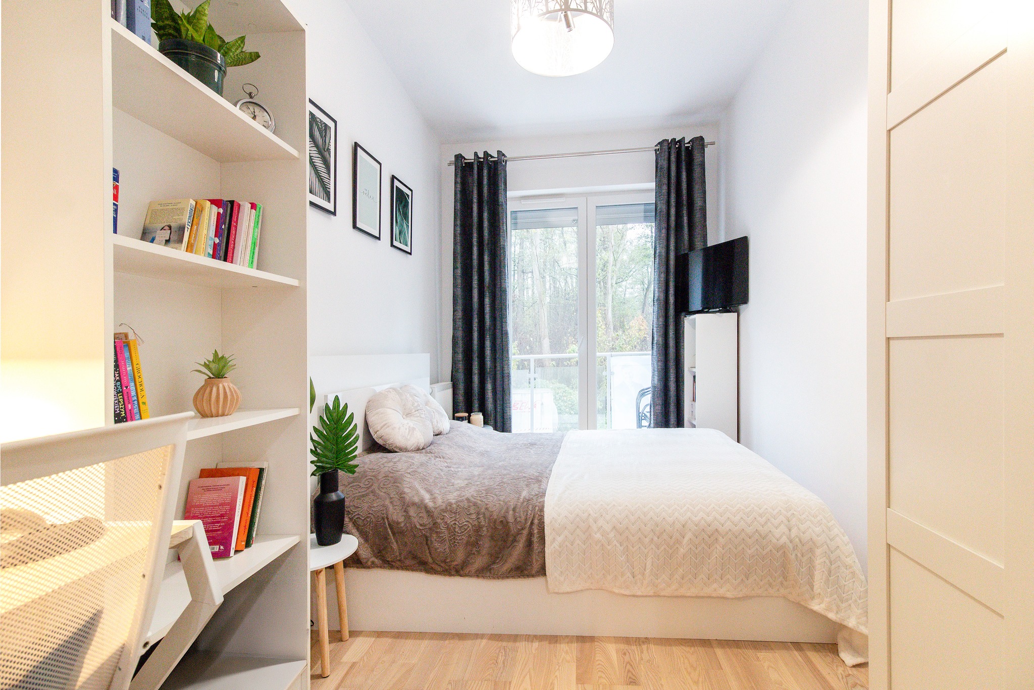 Jasna sypialnia z białymi ścianami, drewnianą podłogą i łóżkiem z dekoracyjną pościelą, widok na balkon przez okno z ciemnymi zasłonami, regał z książkami i dekoracjami po lewej stronie...