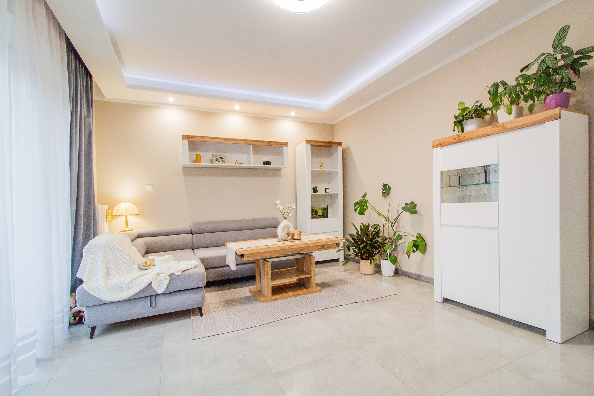 Jasne wnętrze salonu z szarą sofą narożną, drewnianym stolikiem kawowym, białą komodą z roślinami doniczkowymi na górze i szklanymi kieliszkami w środku, oraz zasłony z delikatnym światłem...