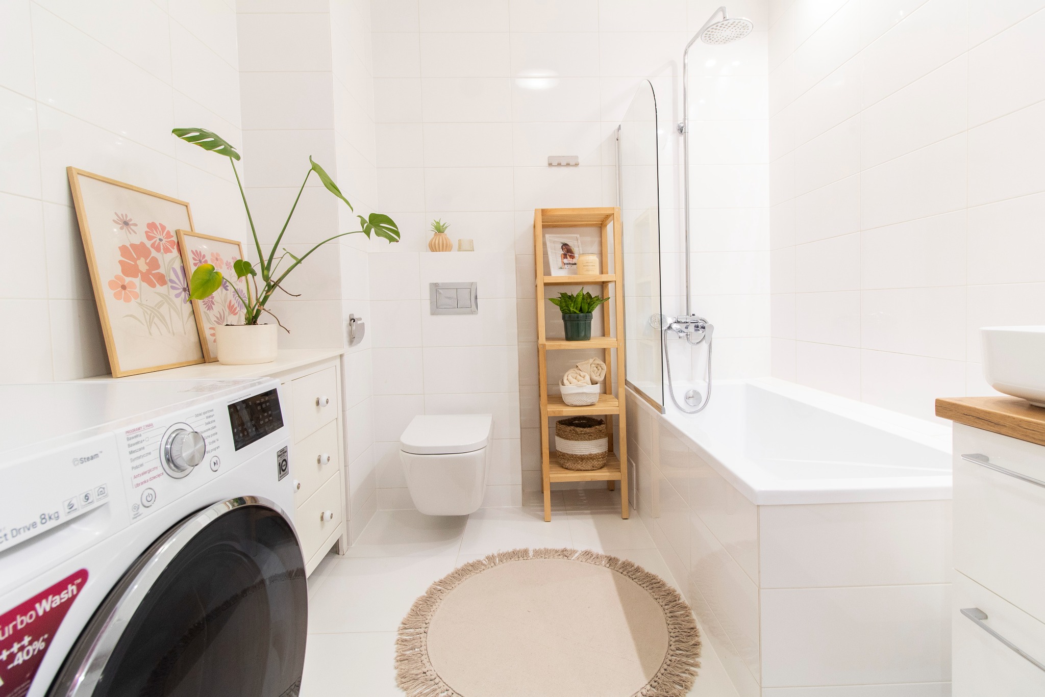 Jasna łazienka w minimalistycznym stylu z białą pralką, toaletą podwieszaną, wanną za szklanym parawanem, drewnianym regałem i okrągłym dywanikiem z frędzlami na białych płytkach podłogowych.