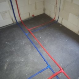 Robert Kulesza - Surowe ściany z betonu komórkowego z poprowadzonymi rurami instalacji wodnej w kolorze czerwonym i niebieskim, ułożonymi na podłodze z ciemnych płytek.