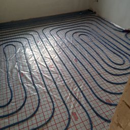 Robert Kulesza - Niebieskie rury ogrzewania podłogowego ułożone na folii z czerwoną siatką, widok z góry, fragment ściany z prawej strony.