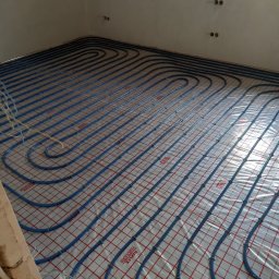 Robert Kulesza - Niebieskie rury systemu ogrzewania podłogowego ułożone na folii z czerwoną siatką w surowym wnętrzu, widoczne kable elektryczne i niedokończona ściana.