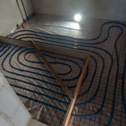 Robert Kulesza - Niedokończona instalacja ogrzewania podłogowego z niebieskimi rurami na folii z czerwoną siatką, tymczasowo zabezpieczona drewnianymi elementami, widok z góry w surowym wnętrzu.