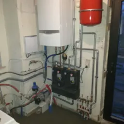 Instalacja centralnego ogrzewania: biały piec gazowy Vaillant, czerwony zbiornik wyrównawczy, czarna skrzynka rozdzielacza z zielonymi diodami, liczne rury miedziane i stalowe, pompa obiegowa...