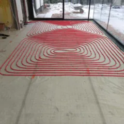 Czerwone rurki systemu rozprowadzenia ciepła, przymocowane zielonymi uchwytami do folii z nadrukowaną siatką, w surowym wnętrzu z widokiem na ośnieżony ogród.