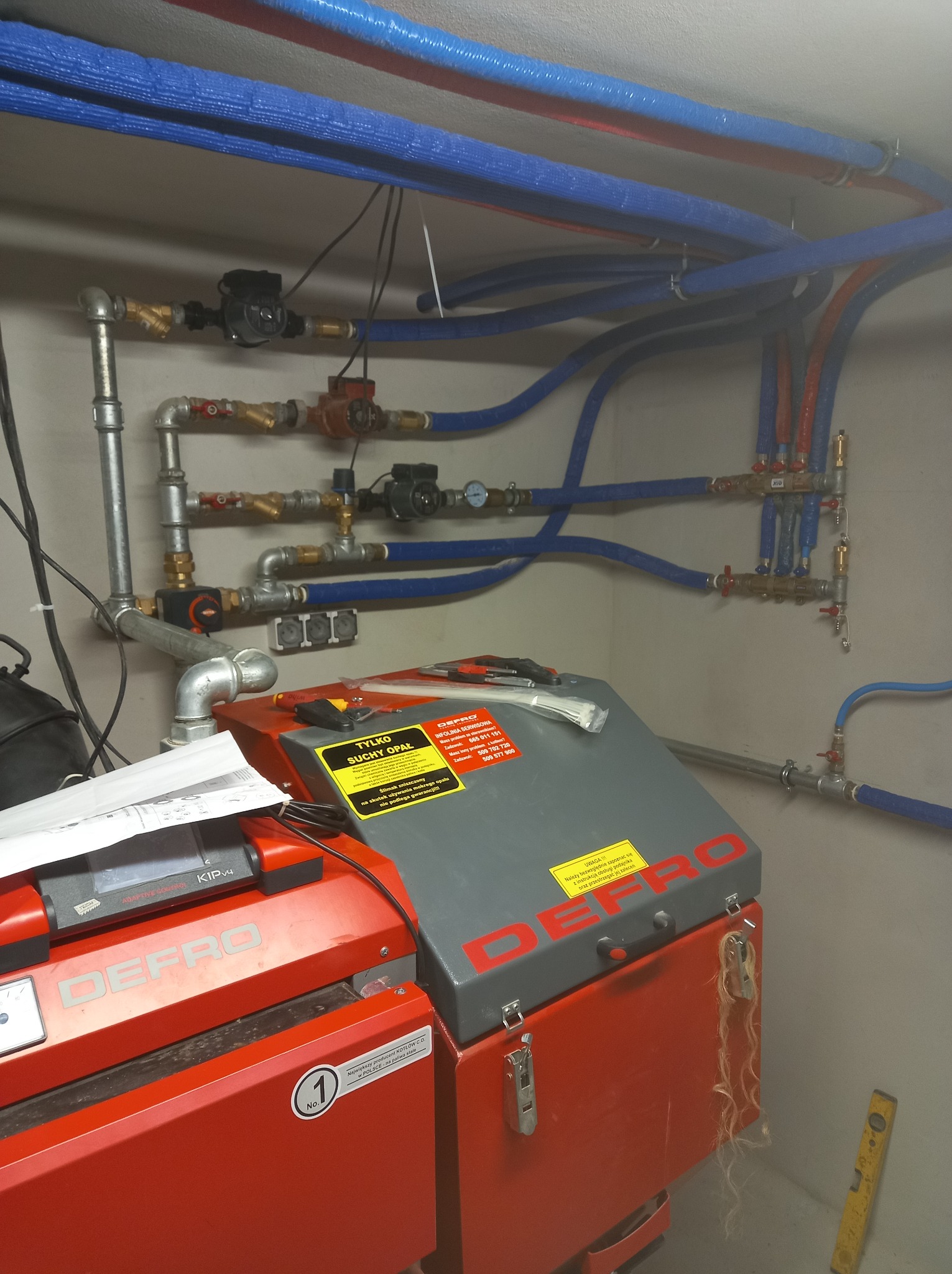 Czerwony piec DEFRO z podłączonymi rurami instalacji grzewczej, izolowane niebieskie rury na ścianie, widoczne zawory i elementy sterujące, poziomica oparta o piec.