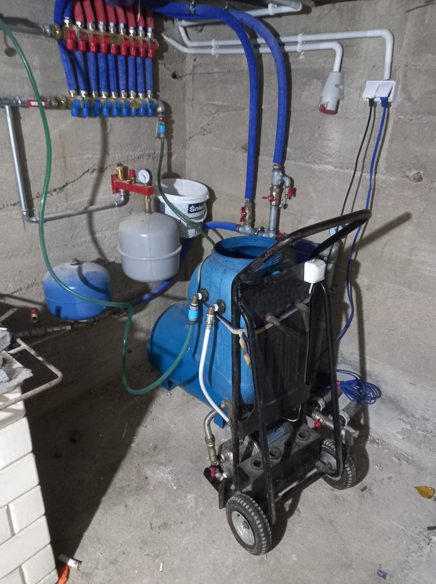 Rozdzielacz instalacji grzewczej z licznymi rurami w kolorach czerwonym i niebieskim, manometr, zbiorniki wyrównawcze i mobilna pompa ciśnieniowa na wózku w piwnicy z surowymi ścianami.