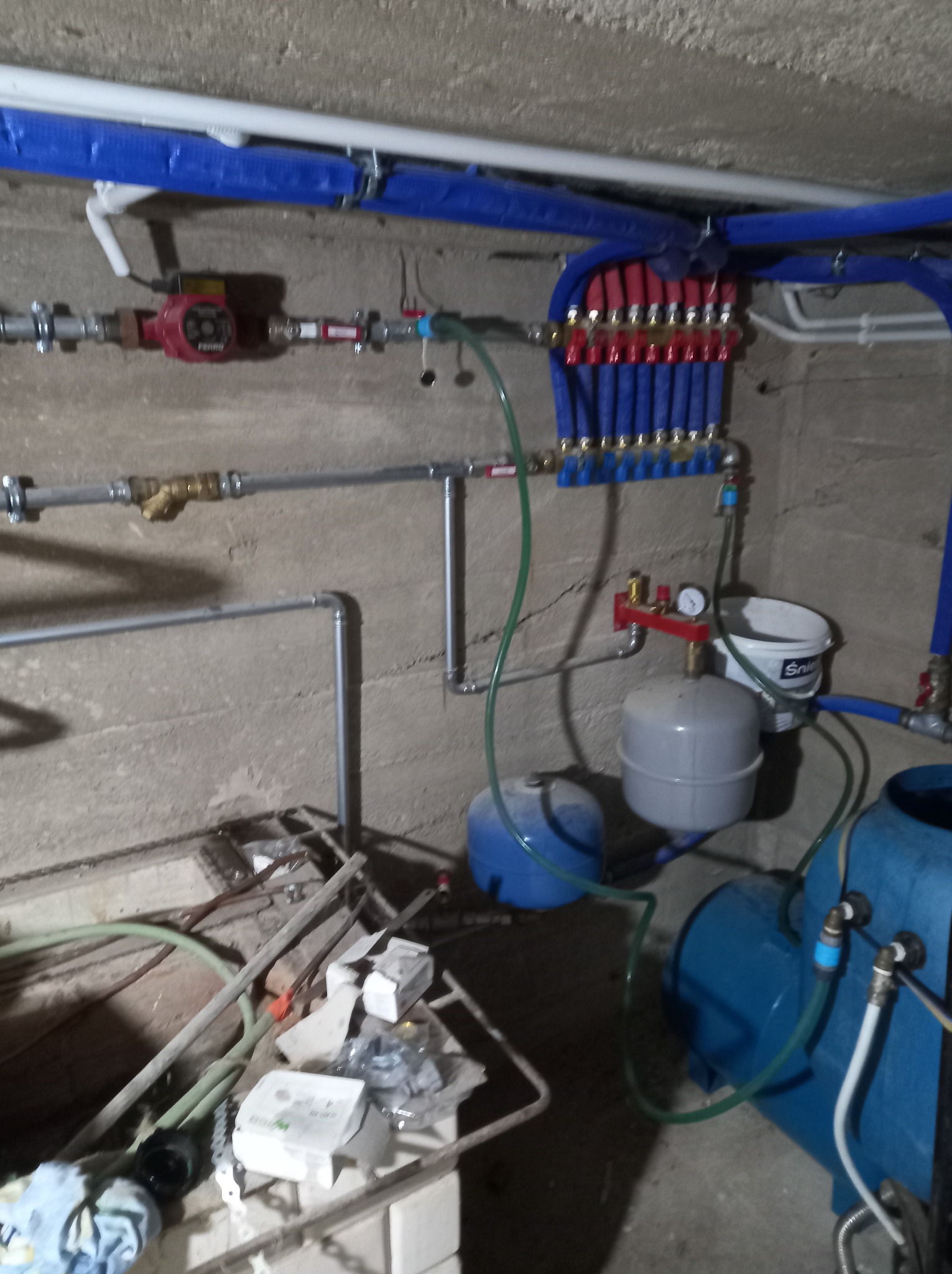 Rozdzielacz instalacji grzewczej z rurami w izolacji, pompą obiegową, manometrem i zbiornikami wyrównawczymi w surowym pomieszczeniu z betonowymi ścianami.