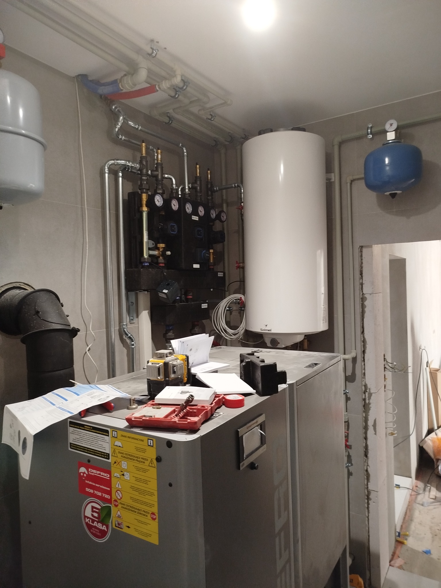 Kotłownia: piec grzewczy Defro z osprzętem, rury instalacji grzewczej pod sufitem, laser krzyżowy i dokumentacja na obudowie pieca.