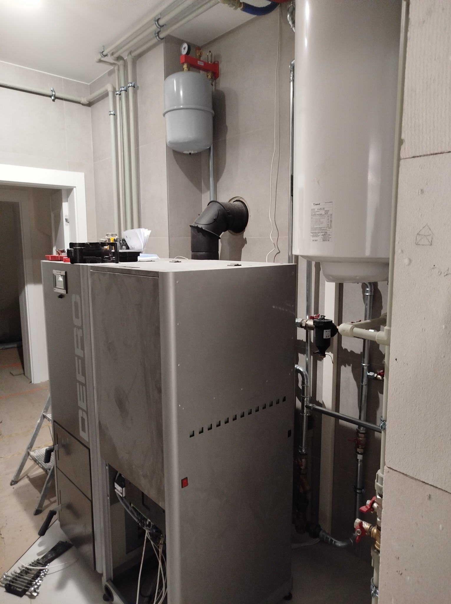 Nowoczesny piec grzewczy Defro w kotłowni, z widocznymi rurami instalacji, zbiornikiem wyrównawczym i narzędziami na podłodze, w trakcie montażu.